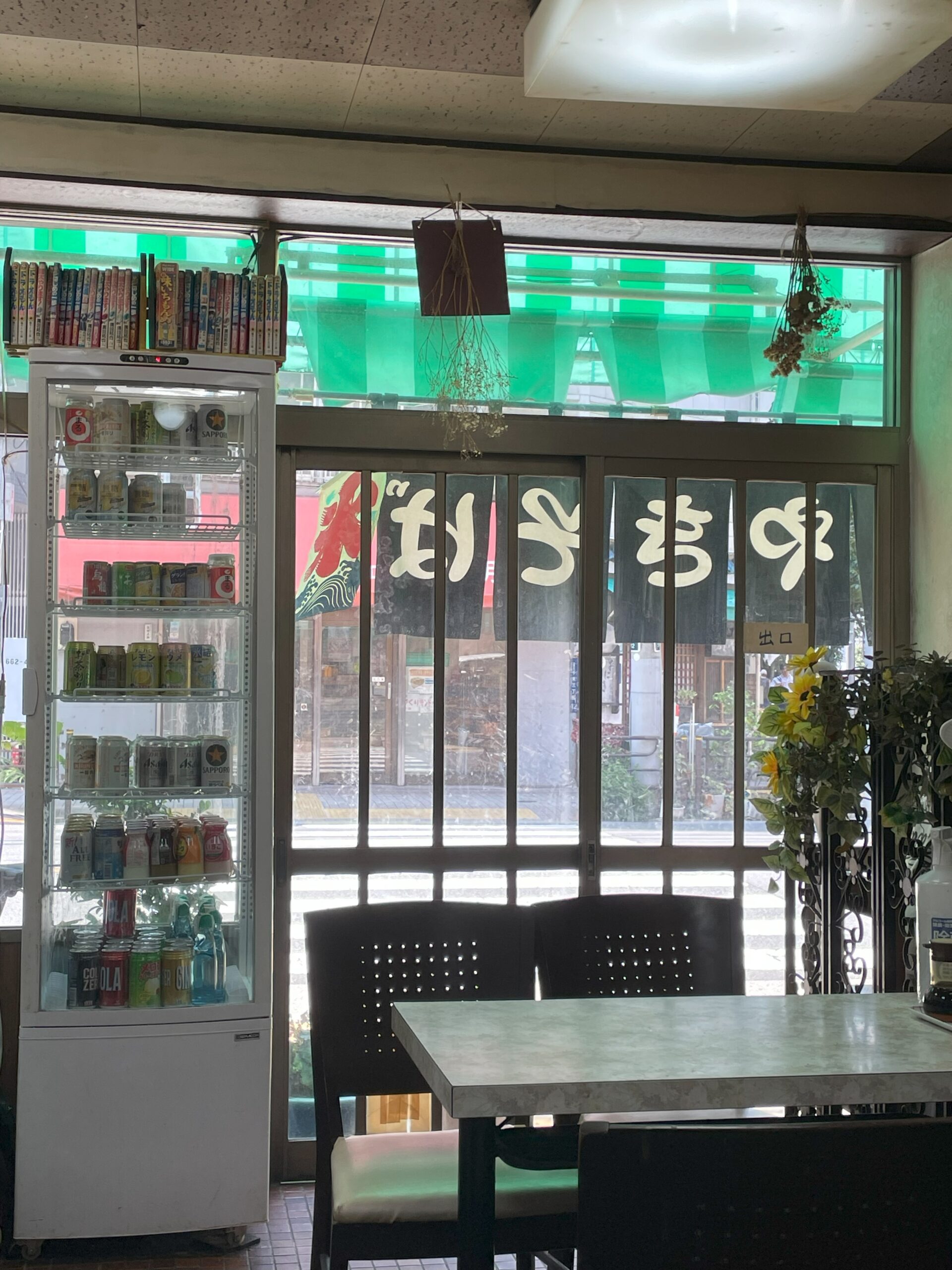 店内の冷蔵庫にはジュースやビールが並び、テーブルと椅子が置かれている。上には漫画本も置かれている。