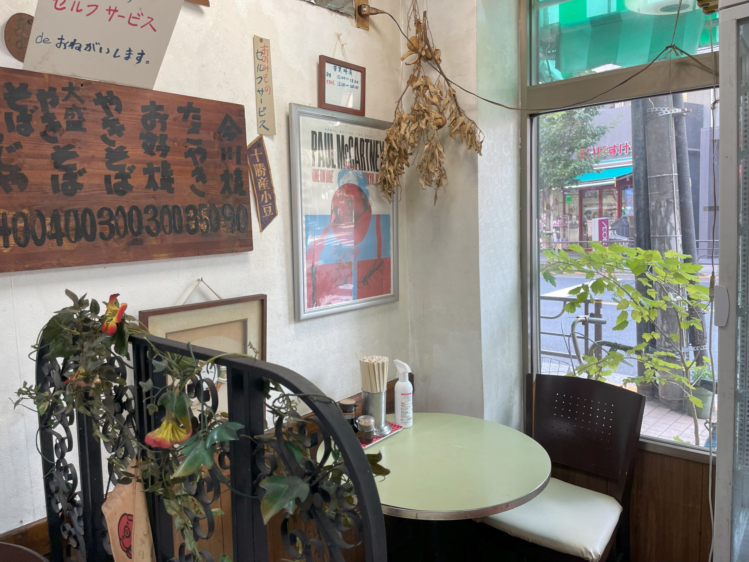 窓際に置かれた丸テーブルと椅子。壁にはポスターや木製メニュー板が飾られている。
