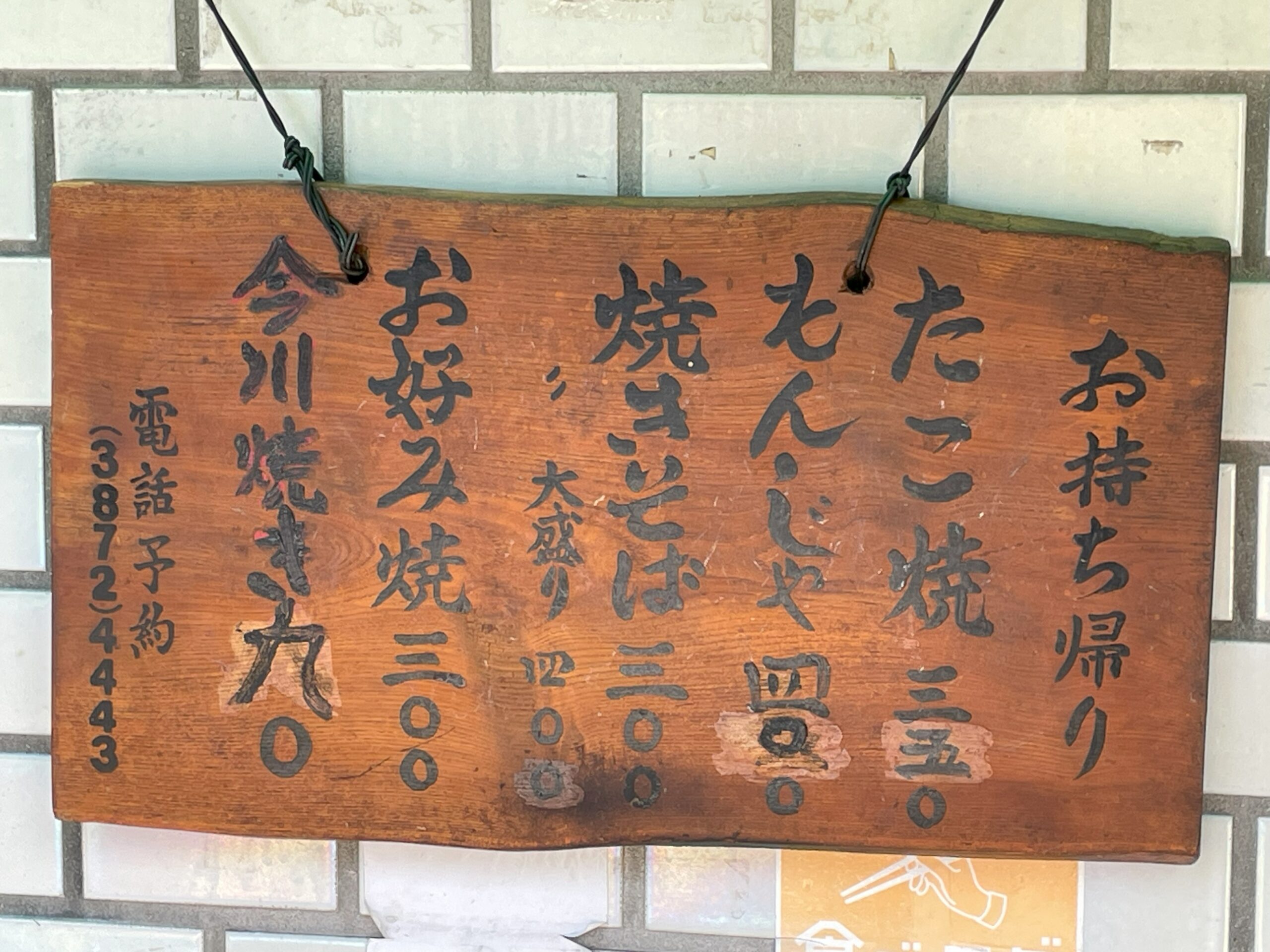 店外の壁に掛けられた木製のメニュー板。たこ焼き、もんじゃ、焼きそば、今川焼などの価格が書かれている。