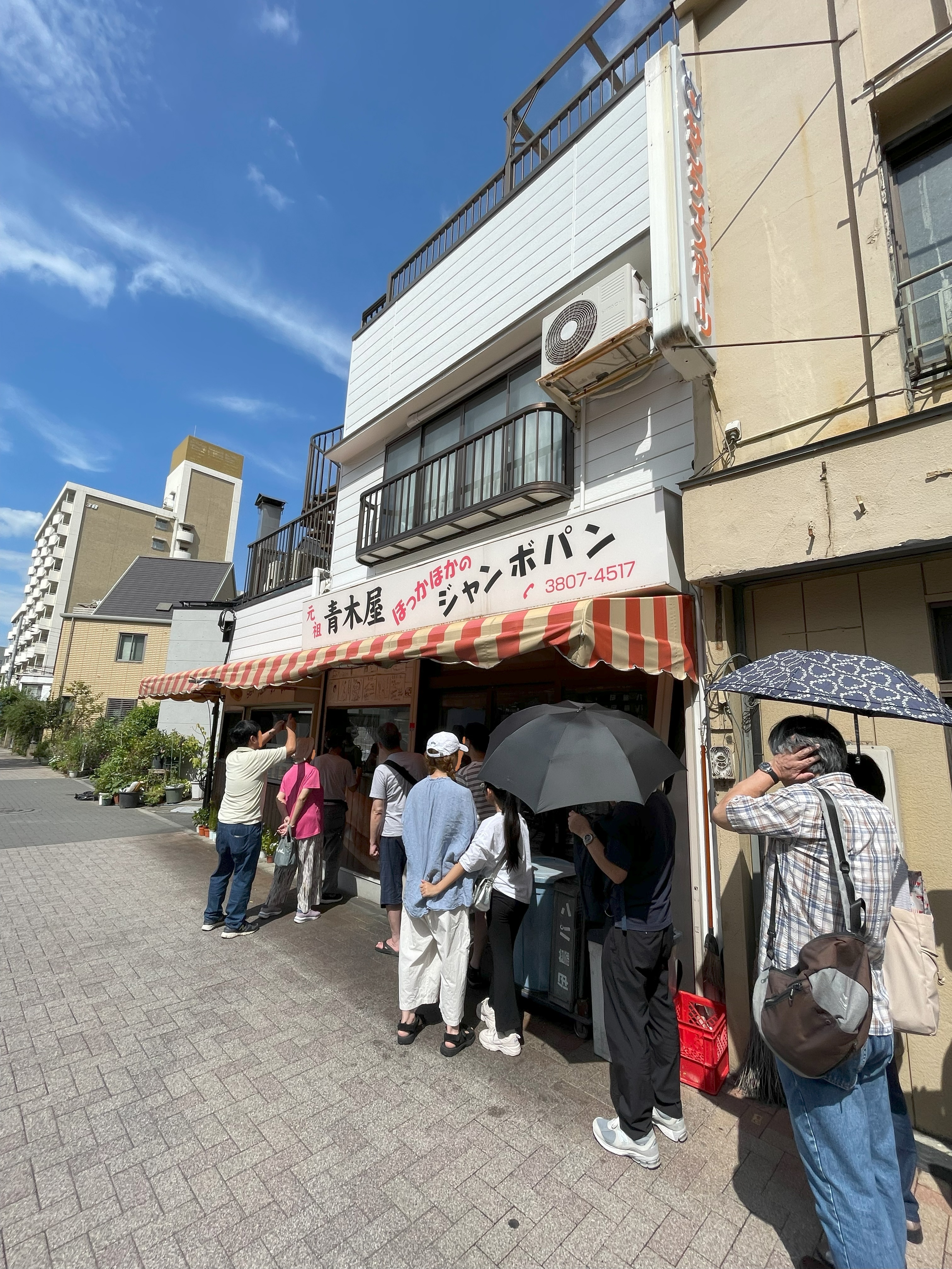 元祖・青木屋さんの店舗横に並ぶ行列。多くの人が日傘をさしてパンの購入を待っている。
