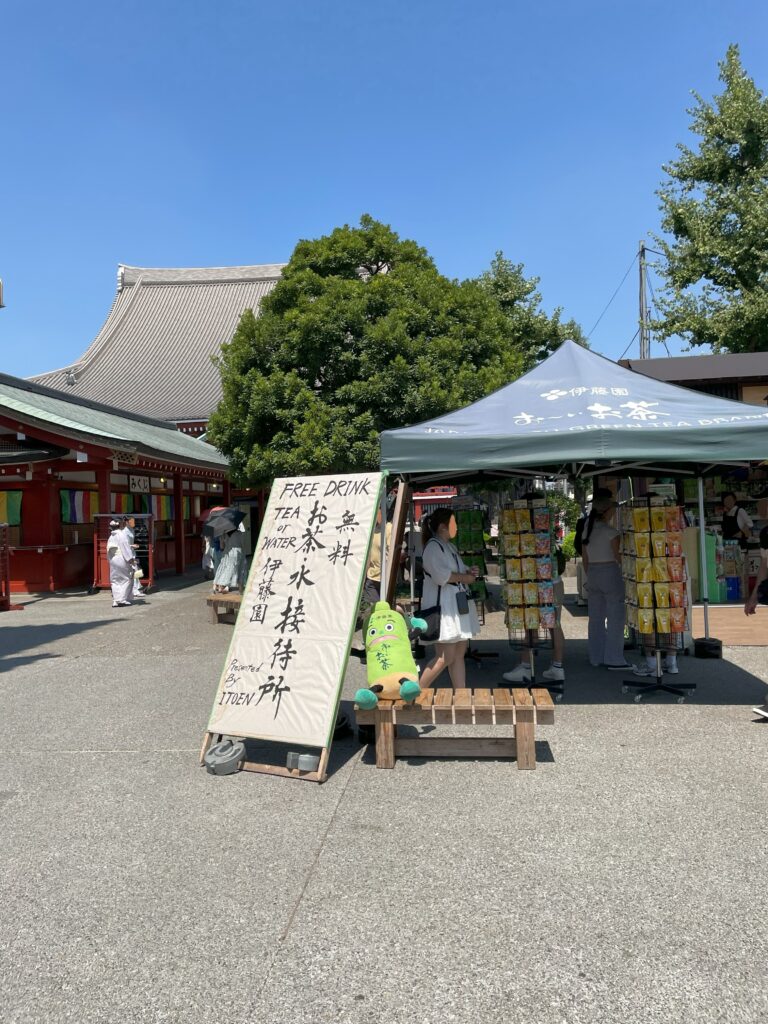 「お茶・水接待所」の看板近くのベンチに「お～いお茶くん」が座っている。