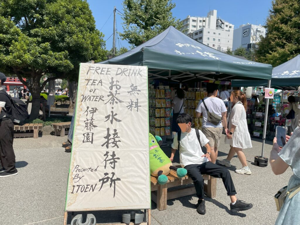 「お茶・水接待所」のベンチに座る「お～いお茶くん」と一緒に記念撮影をする観光客の様子。