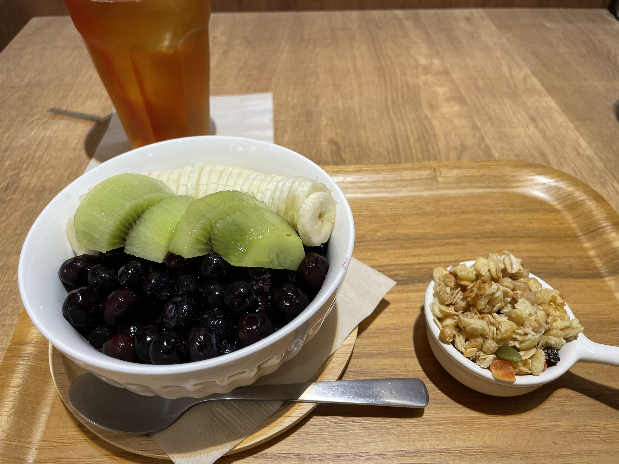 アサイーボウルの上にキウイ、バナナ、ブルーベリーが彩りよく盛りつけられ、横にグラノーラとアイスティーが添えられている。