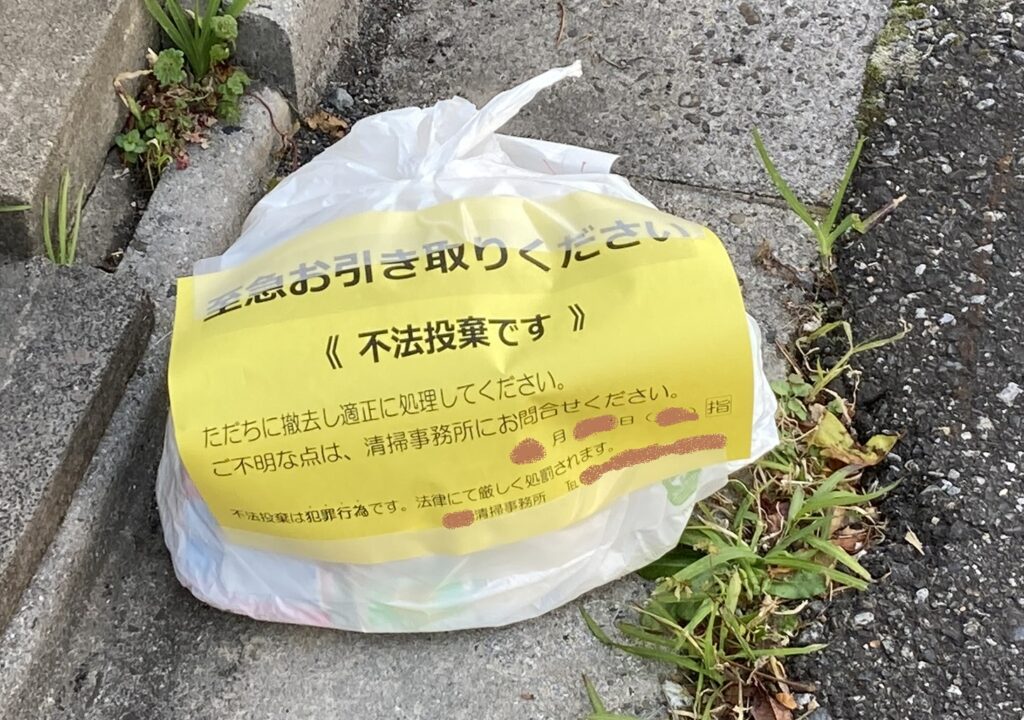 路上に置かれた不法投棄のゴミ袋に黄色い注意シールが貼られている様子。