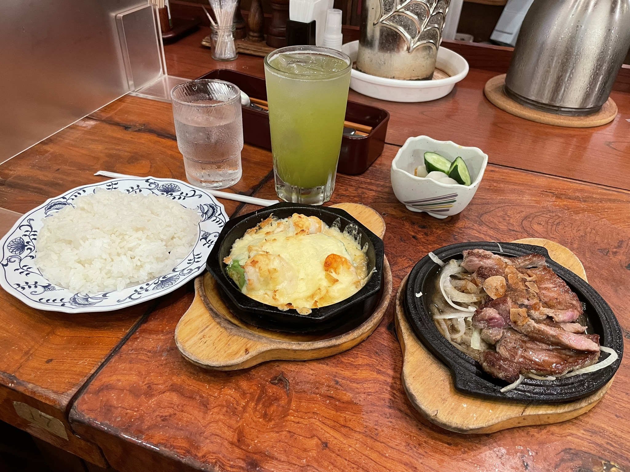 鉄皿で提供される「ミニサーロウィンステーキ」「えびとアボカドのチーズ焼き」「アイス抹茶（ソフトドリンク）」のセットと、ご飯とお新香。