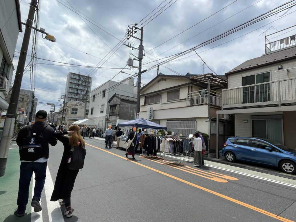 曇り空のもと、露店と行き交う人々。衣料品のハンガーラックが路上に並び、近隣住民や遠方からの来場者が思い思いに品を手に取る。