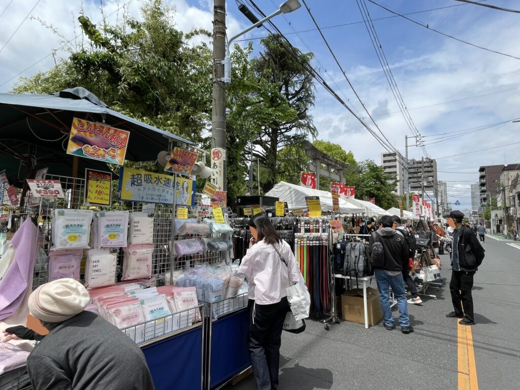 「超吸水速乾ヘアドライタオル」「こんこん祭りだよ」の賑やかな幟が並ぶタオル・日用品ブース。奥にはテントが連なり、境内周辺一帯に広がる市の活気が感じられる。