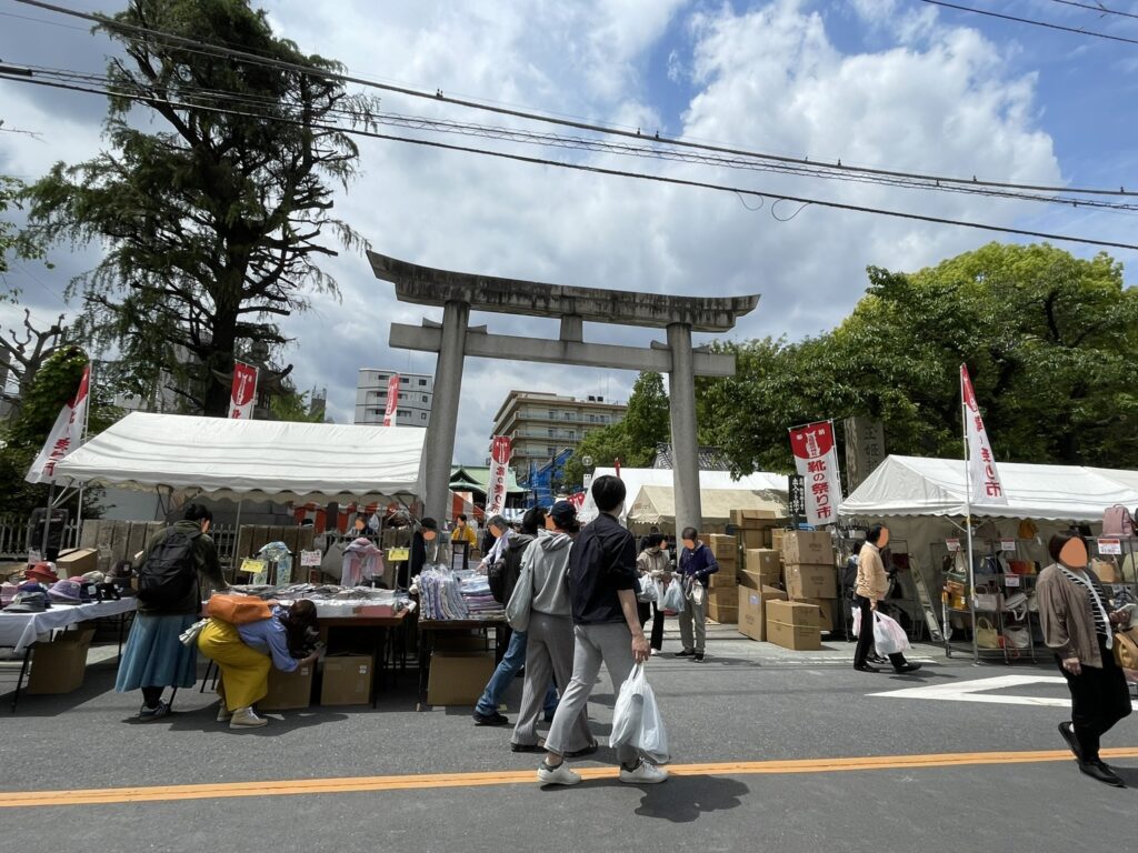 玉姫稲荷神社の大きな鳥居を背景に「靴の祭り市」の幟旗がたなびく。境内の入口にも出店が並び、神社と市が一体となった「こんこん靴市」ならではの光景。