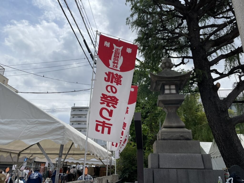 境内の燈籠の傍らにはためく「靴の祭り市」の大幟。稲荷神社の使いであるお狐様の絵が描かれ、「こんこん」の名の由来を静かに物語っている。