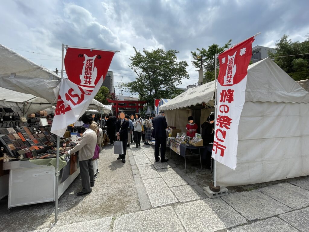 「靴の祭り市」の幟がはためく参道で、革小物を手に取る来場者の姿。奥には朱色の鳥居が見え、境内全体がお祭りの雰囲気に包まれている。