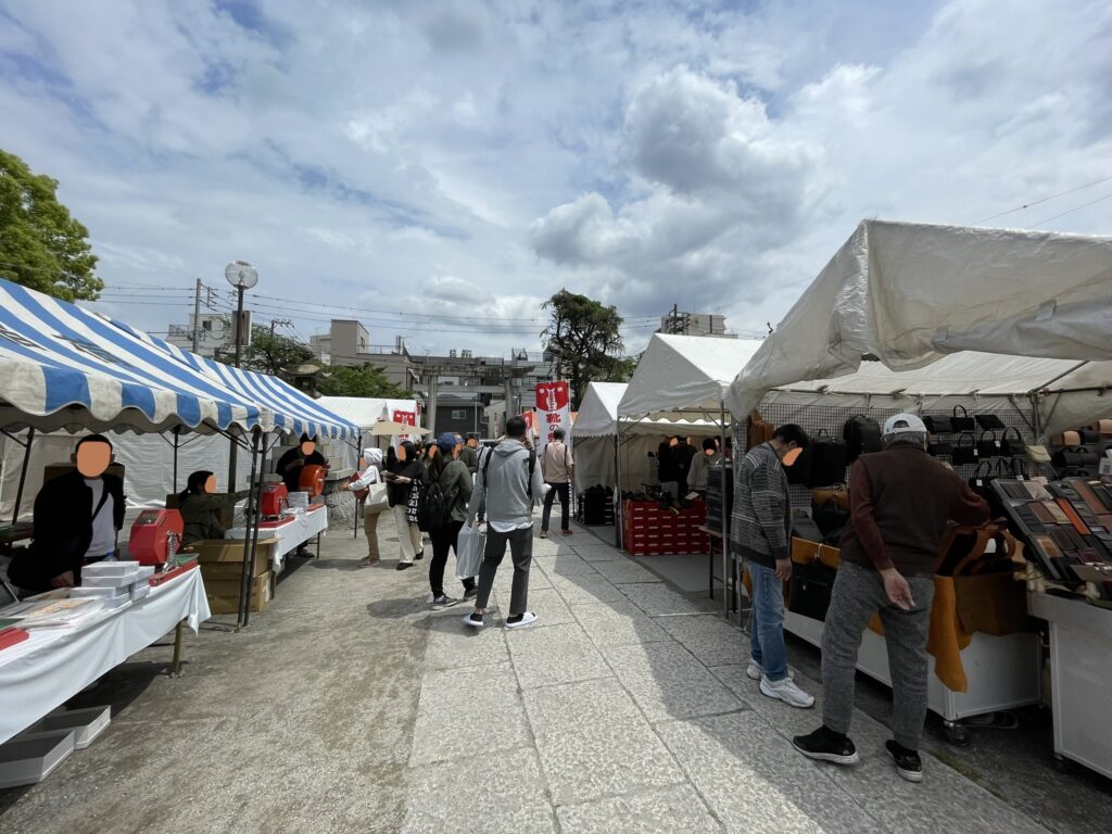 白と青のテントが立ち並ぶ境内の広場。革製品や靴のブースが左右に続き、大鳥居を背景ににぎわう「こんこん靴市」の会場風景。