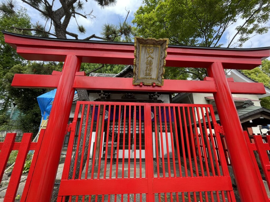 「口入稲荷社」と刻まれた扁額を掲げる朱色の鳥居。格子の扉の向こうに社殿が見え、長い歴史を持つ玉姫稲荷神社の境内に息づく信仰の静けさが漂う。