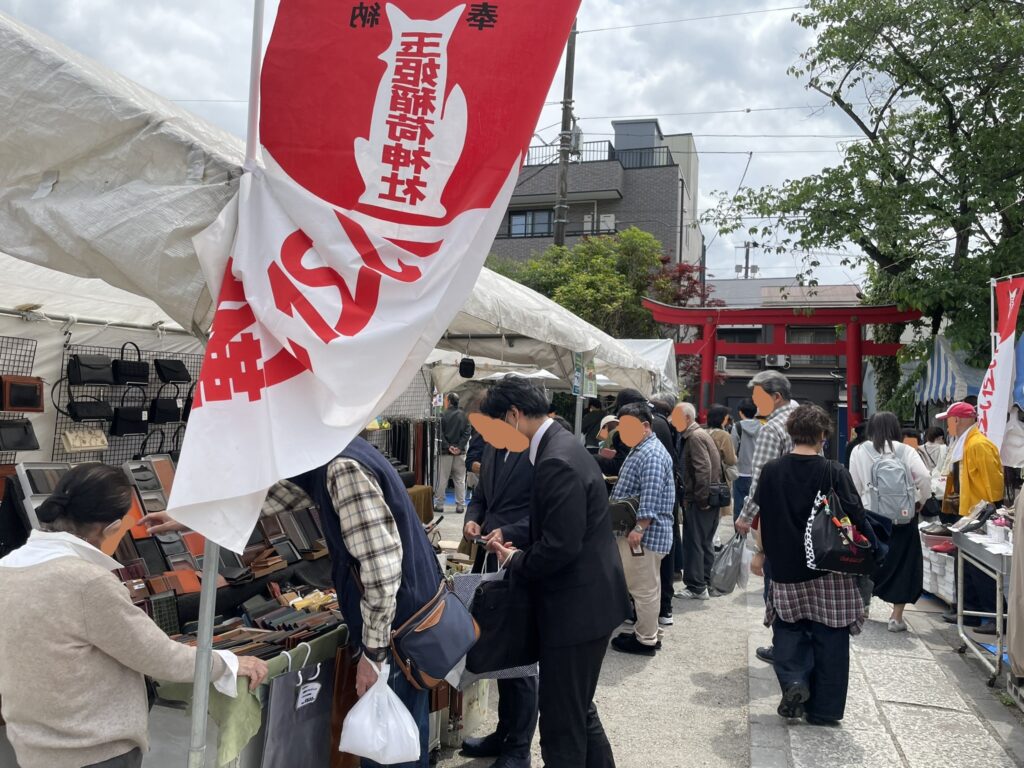お狐様が描かれた「靴の祭り市」の幟を前景に、革小物のブースと来場者が行き交う賑やかな参道。奥には朱色の鳥居が見え、神社と市が一体となった光景が広がる。