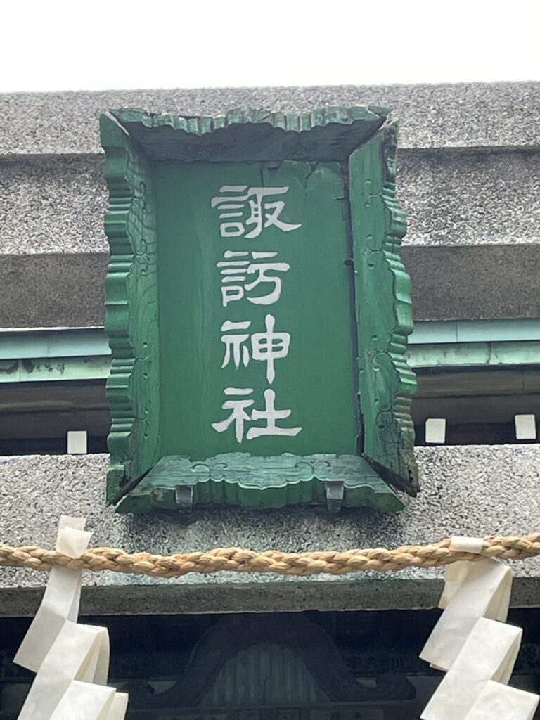 「諏訪神社」と書かれた緑色の社号額（扁額）のアップ。