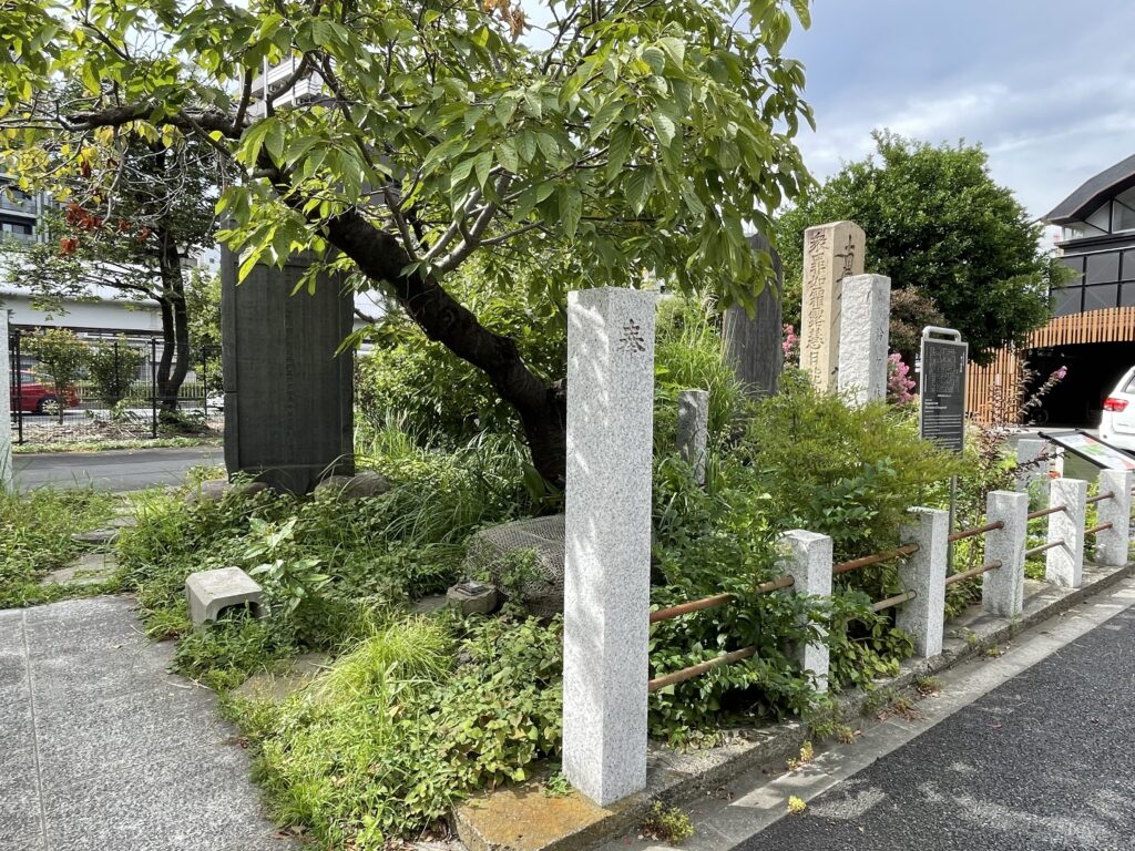 手前は「奉」と刻まれた石柱。奥に見える題目塔の側面には「衆罪如霜露 慧日能消除（多くの罪は霜や露のようなもので、仏の智慧の光で消し去ることができる）」という経文が刻まれている。草むらの中に金網をかぶせた石組みがあり、首洗いの井戸と伝わるものとも見える