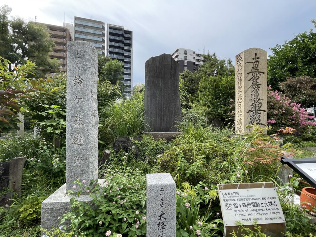 左は「都旧跡 鈴ヶ森遺跡」の石柱、右は「南無妙法蓮華経」の髭題目を刻んだ題目塔。手前には「二十五番 大経寺」の標柱と、しながわ百景「鈴ヶ森刑場跡と大経寺」の案内板が立っている。しながわ百景は昭和62年（1987年）に選ばれた