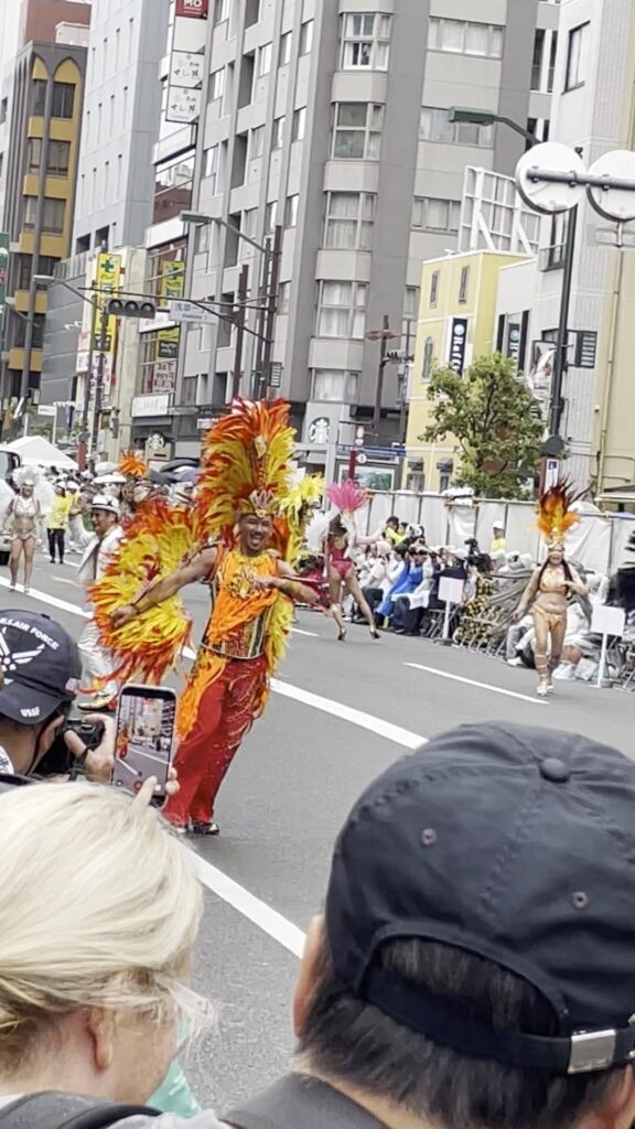 羽根飾りの衣装をまとったダンサーが、沿道に笑顔を向けます。